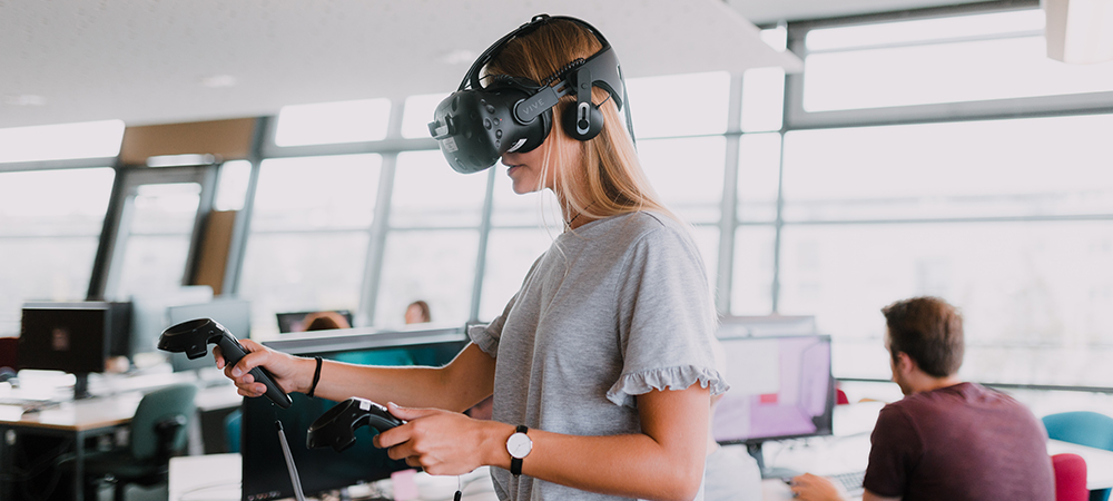 Eine Studentin steht im Games-Lab und probiert ein VR-Spiel aus. Sie trägt dazu eine VR-Brille und hat 2 Sensoren in der Hand, die Ihre Hand- und Arm-Bewegungen in das Spiel integrieren.