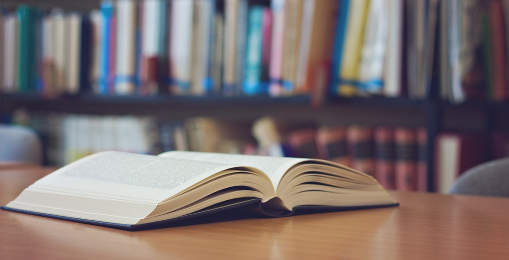 Symbol für Bibilothek und Lernwelt. Das Bild zeigt ein Buch welches aufgeklappt auf einem Tisch in einer Bibliothek liegt.