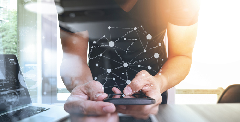 Symbol für Content-Management-Systeme. Das Bild zeigt Hände, die ein Handy halten. Die Hände befinden sich mittig auf dem Bild. Links ist ein Laptop zu erkennen.
