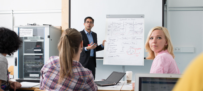 A photo of a teacher in front of some students. The teacher is next to a whiteboard covered in writing.