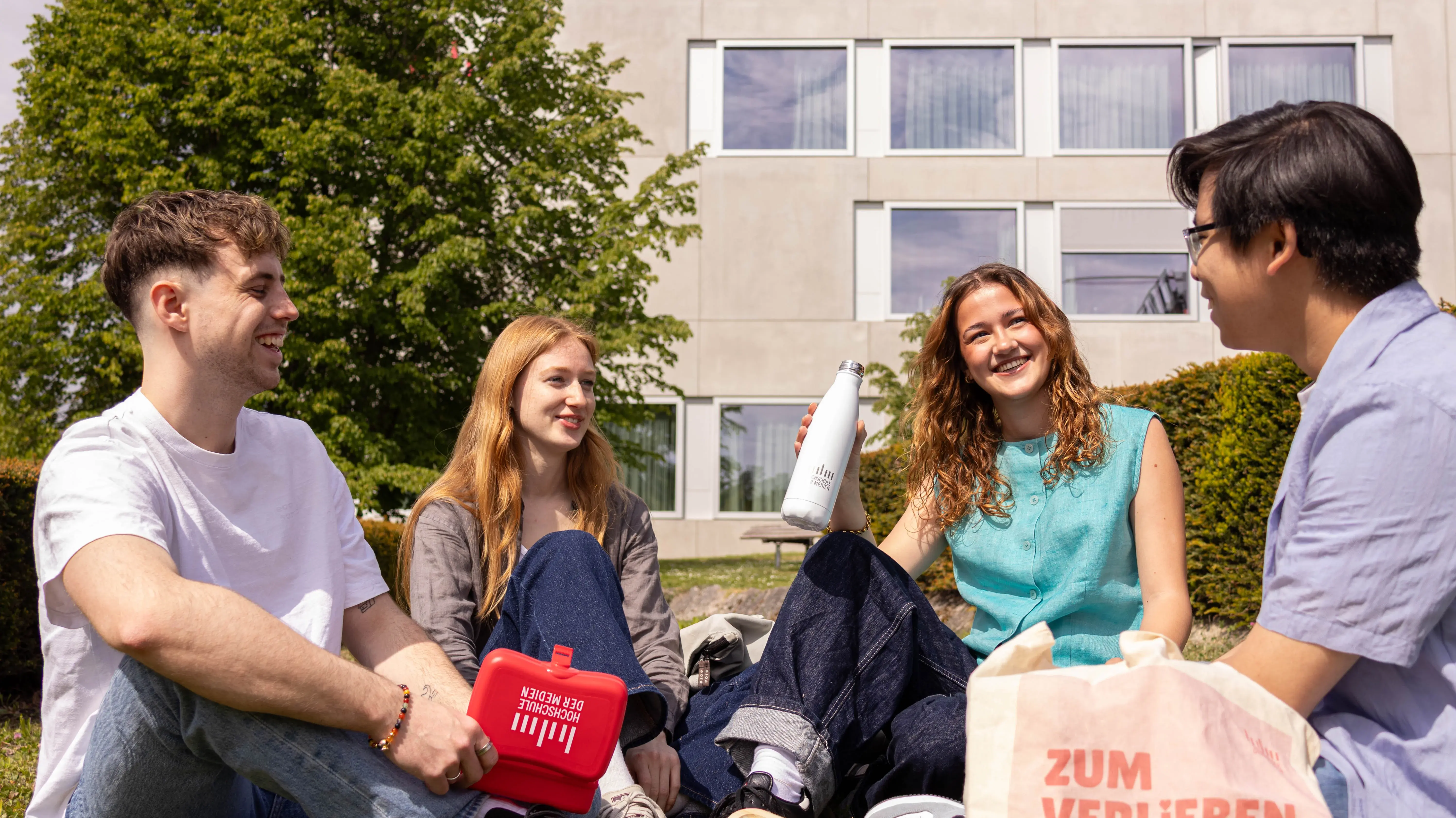 Students spending their break on the lawn of the sunny campus of the Hochschule der Medien.