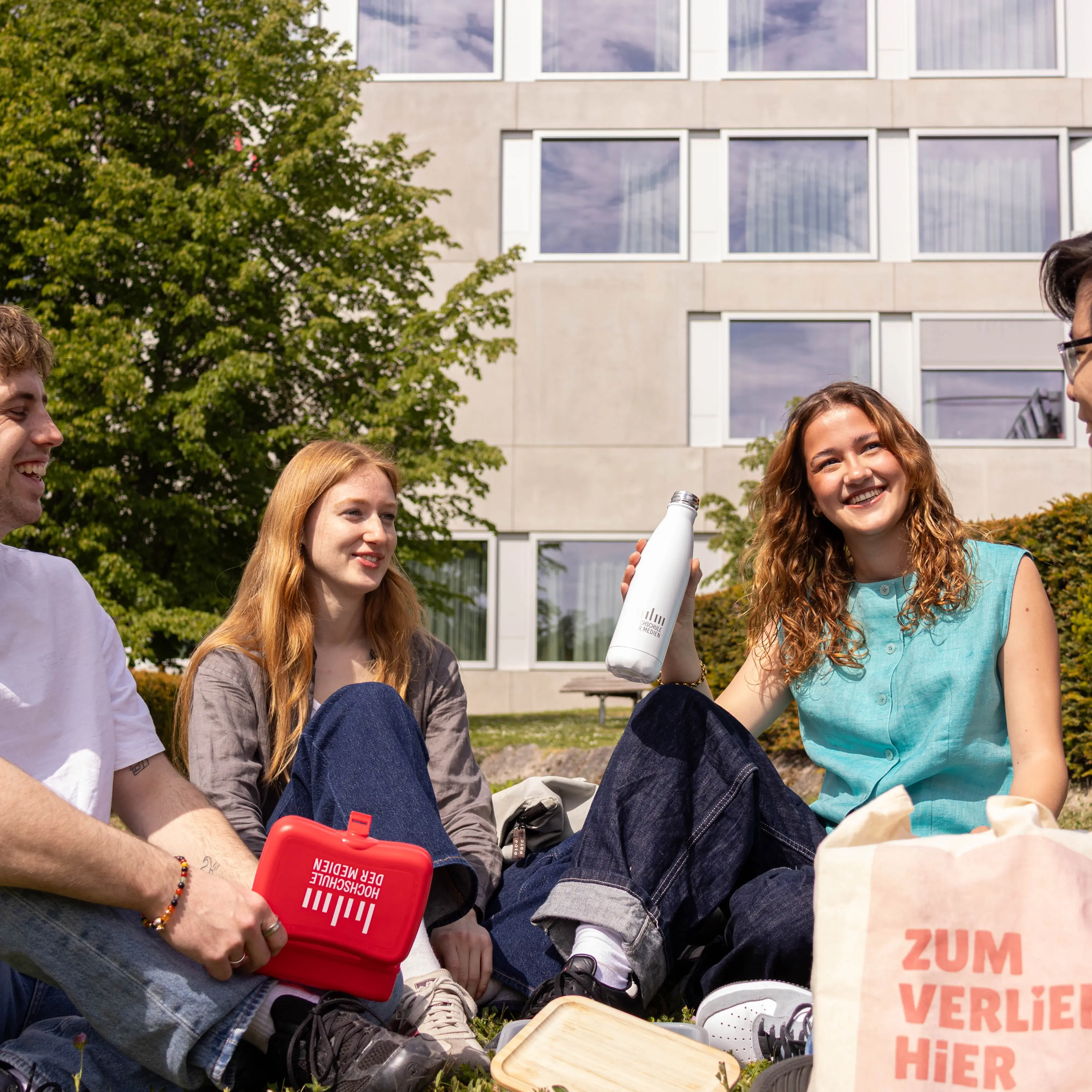 Students spending their break on the lawn of the sunny campus of the Hochschule der Medien.