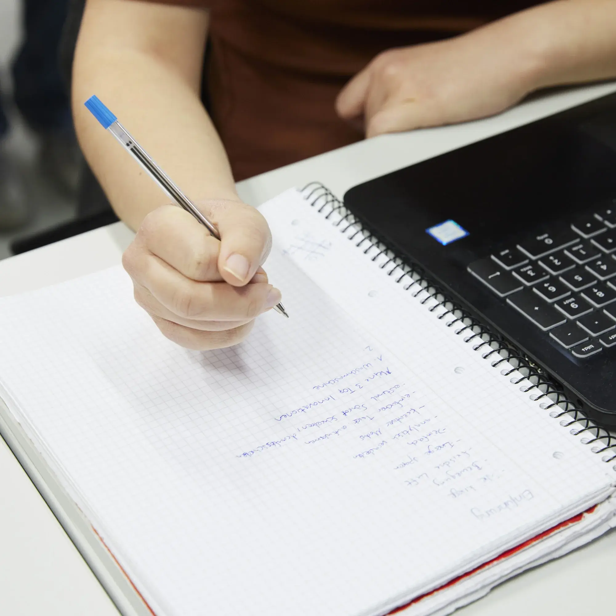 Someone is jotting down information on a notepad next to their laptop.
