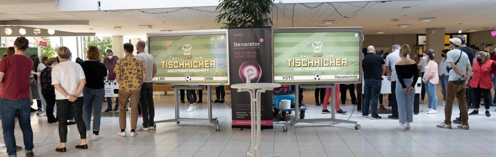 Bild der 6. HdM Tischkicker WM. Es sind zwei Bildschirme mit den laufenden Partien und die Besucher sowie Spieler*innen zu sehen.